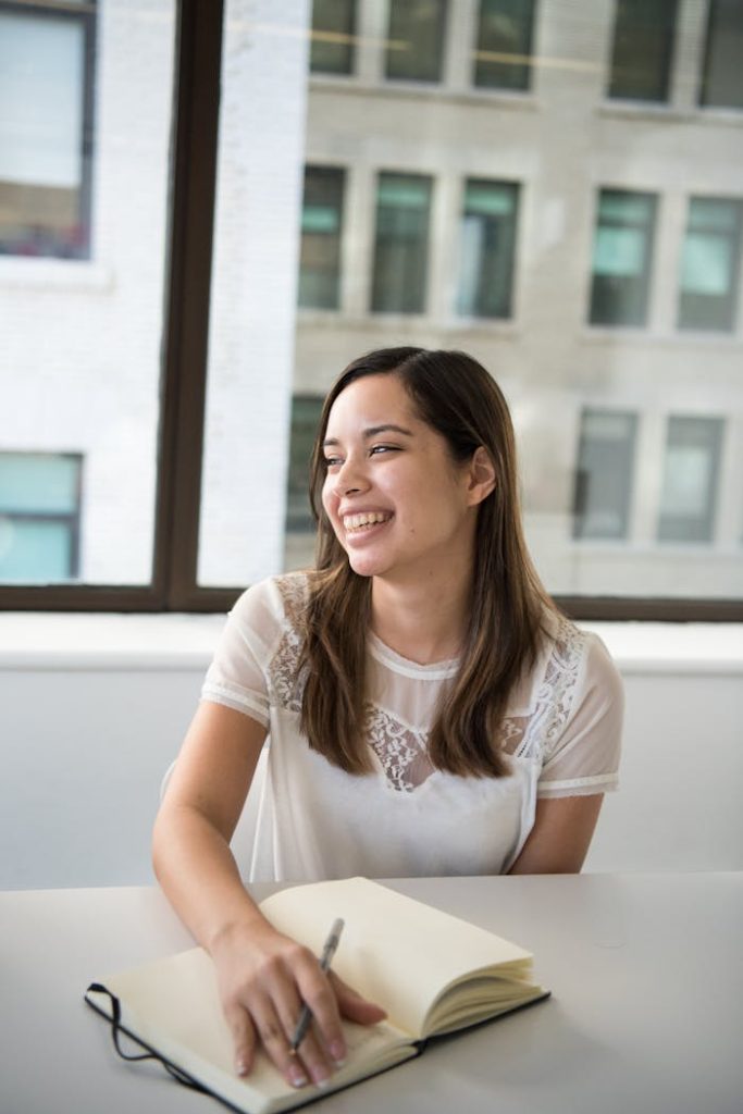 Woman enjoying writing in a notebook with natural daylight in modern office.